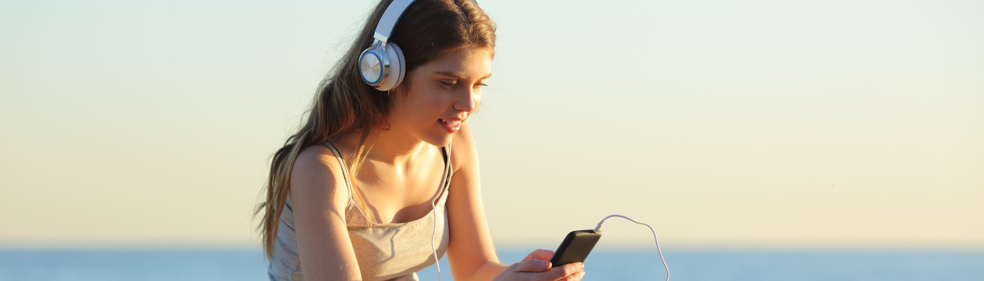 Mädchen mit Headphones schaut schaut entspannt aufs Handy und sitzt am Meer