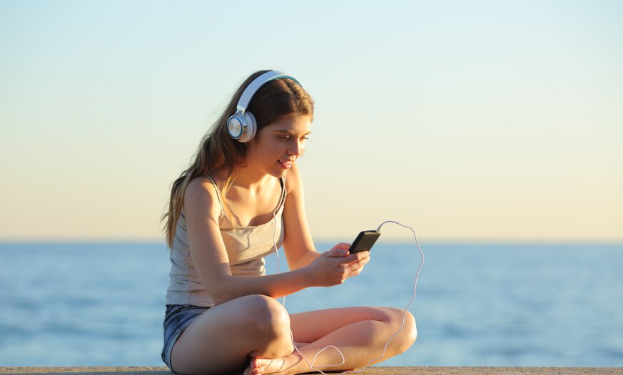 Mädchen mit Headphones schaut schaut entspannt aufs Handy und sitzt am Meer