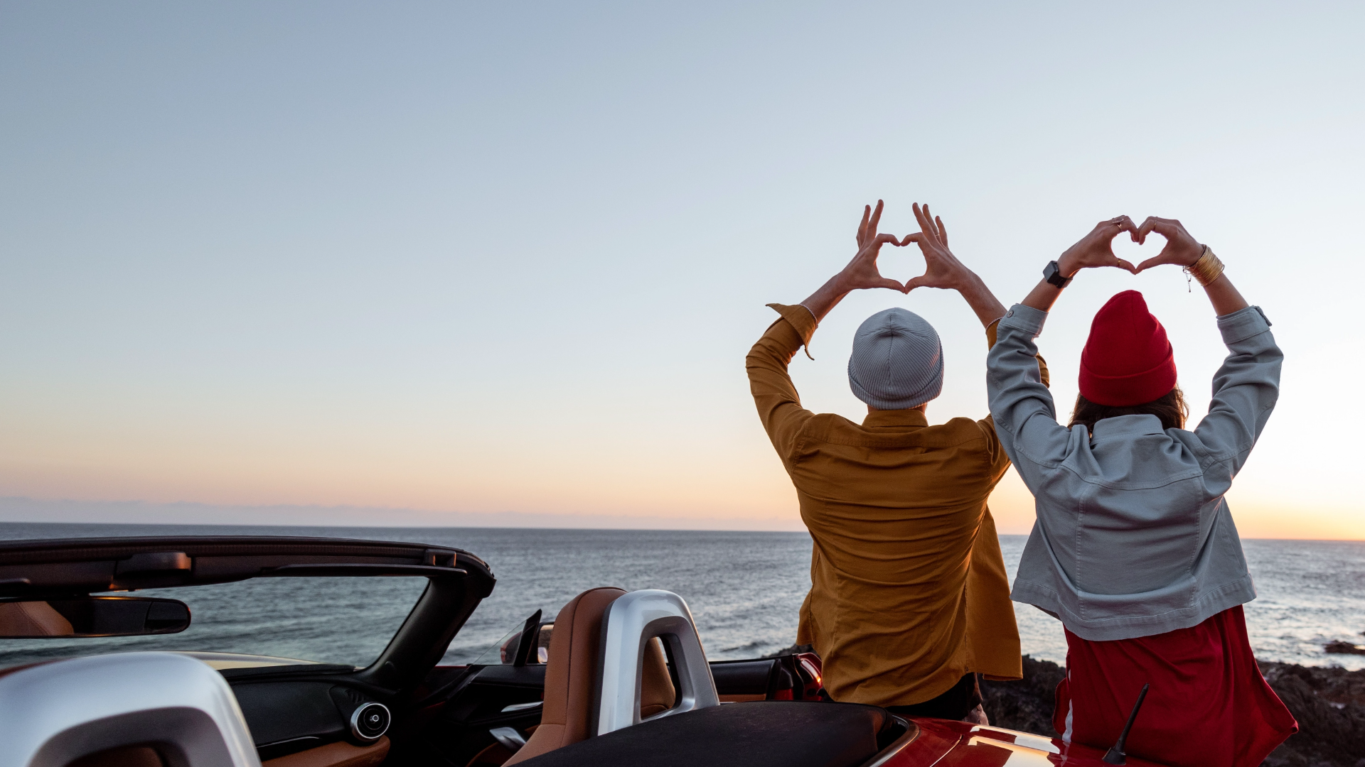Mädchen und Junge von hinten sitzen auf Auto bei Sonnenuntergang und formen Herz mit Händen